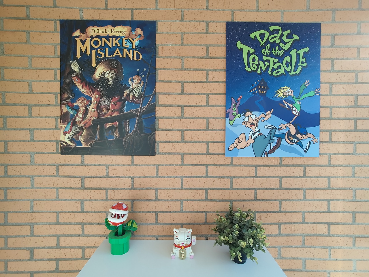 Pósters de videojuegos retro Monkey Island y Day of the Tentacle en pared de ladrillo rojo, con adornos.