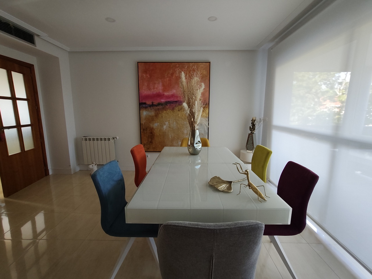 Comedor moderno con sillas coloridas y mesa blanca, decorado con jarrón y cuadro abstracto en pared blanca.