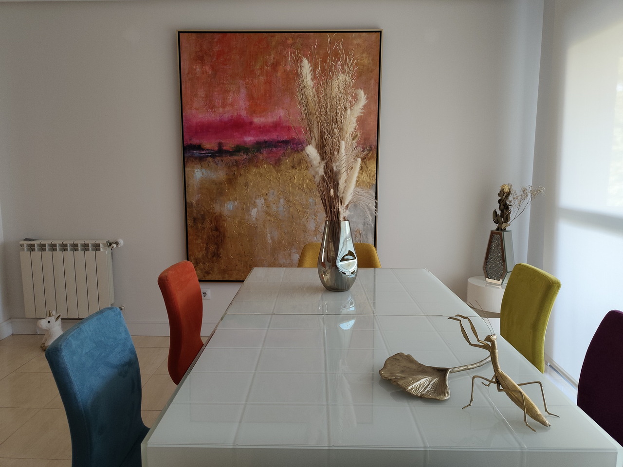 Comedor moderno con sillas de colores, mesa blanca, jarrón plateado y cuadro abstracto en la pared.