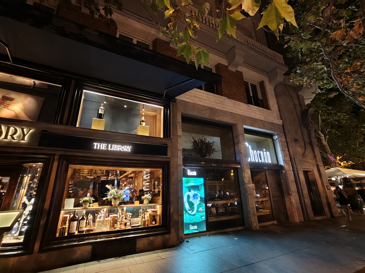 Fachada de tiendas iluminadas por la noche en una calle arbolada, exhibiendo botellas y productos en vitrinas atractivas.