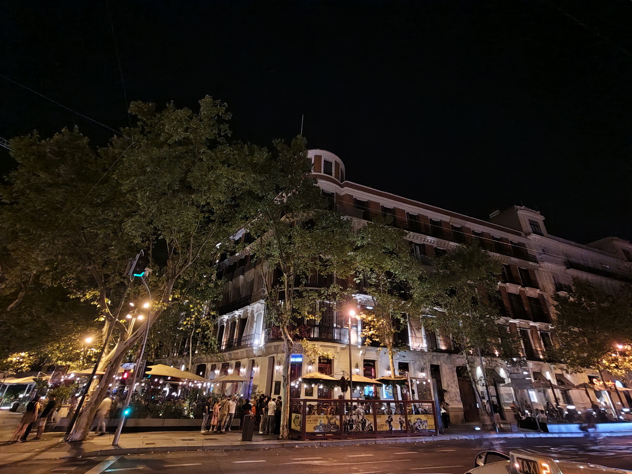Edificio iluminado por la noche con terrazas concurridas, árboles y luces en una calle urbana.