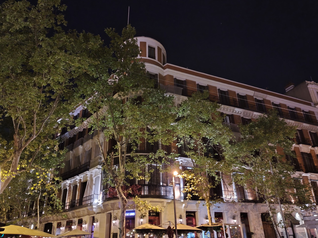 Edificio antiguo iluminado por la noche, rodeado de árboles en una calle urbana con tiendas y luces cálidas.