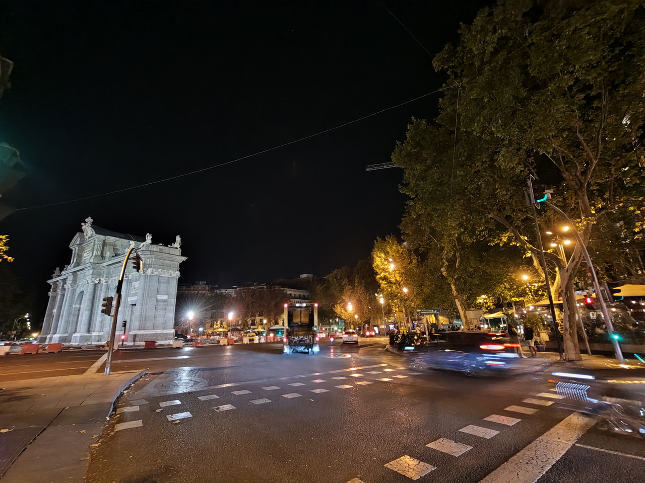 Cruzando la Puerta de Alcalá iluminada por la noche, vista desde una concurrida intersección en Madrid.