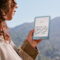 Mujer leyendo un libro digital en un dispositivo Kindle azul con paisaje montañoso de fondo.