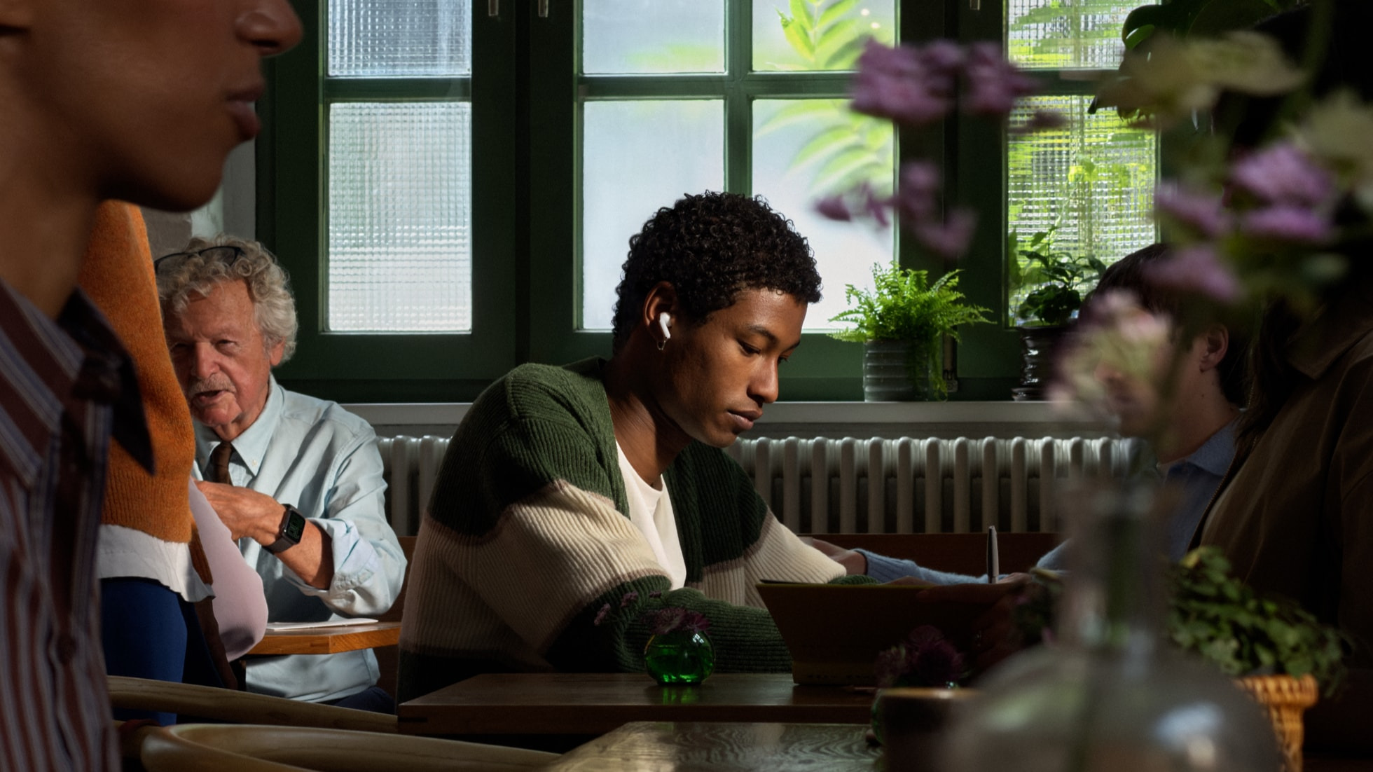 Joven estudiando con audífonos en cafetería iluminada, rodeado de plantas y personas conversando.