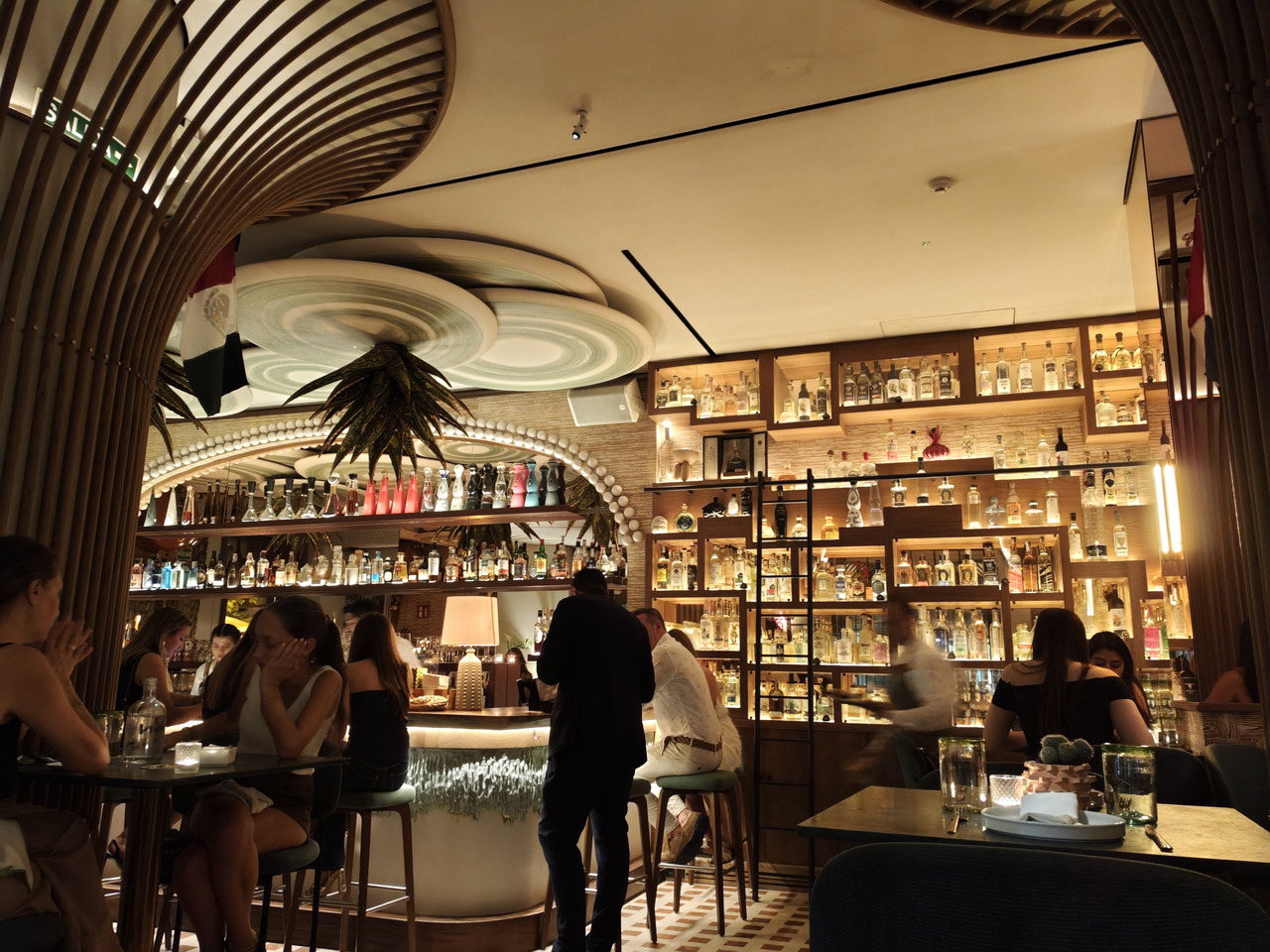Bar moderno y elegante con estanterías repletas de botellas iluminadas, clientes charlando y ambiente acogedor.