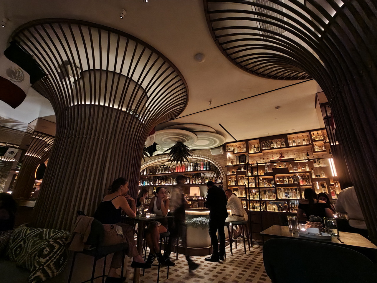 Bar con iluminación ambiental, estanterías llenas de botellas y clientes disfrutando de una noche en un espacio moderno.