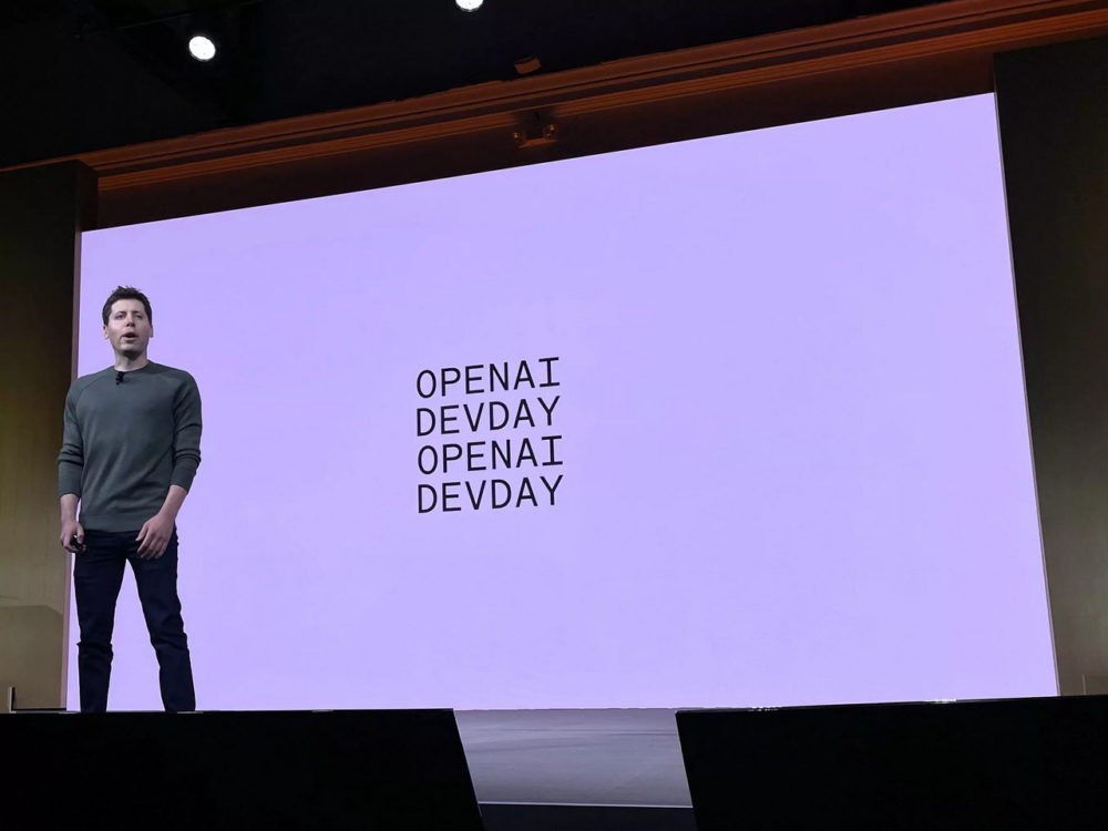 Persona hablando en el evento de OpenAI DevDay, con fondo blanco y texto en pantalla.