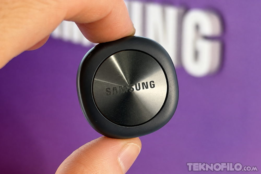 Botón inteligente Samsung en primer plano sobre fondo morado, sostenido con dos dedos.