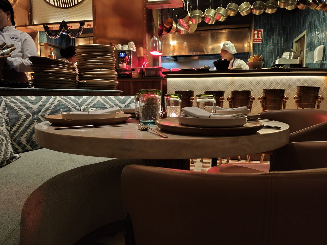 Mesa en restaurante elegante con platos y cubiertos, cocina abierta y chefs al fondo, ambiente acogedor.