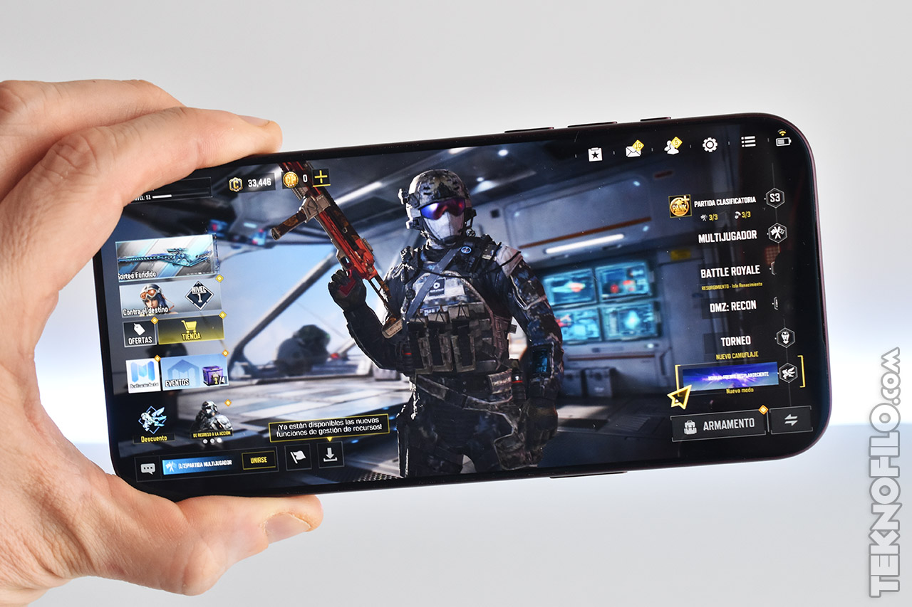 Mano sosteniendo smartphone con juego de batalla en pantalla, mostrando personaje militar equipado y menú de opciones.
