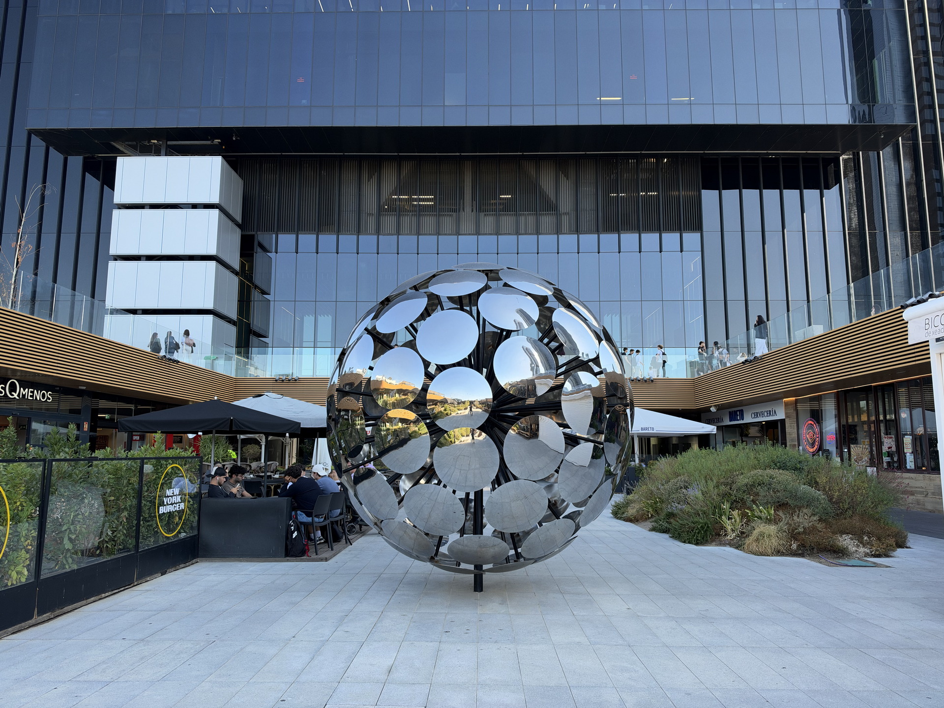Escultura esférica de metal reflejante frente a un edificio moderno de vidrio y tiendas en un centro comercial.