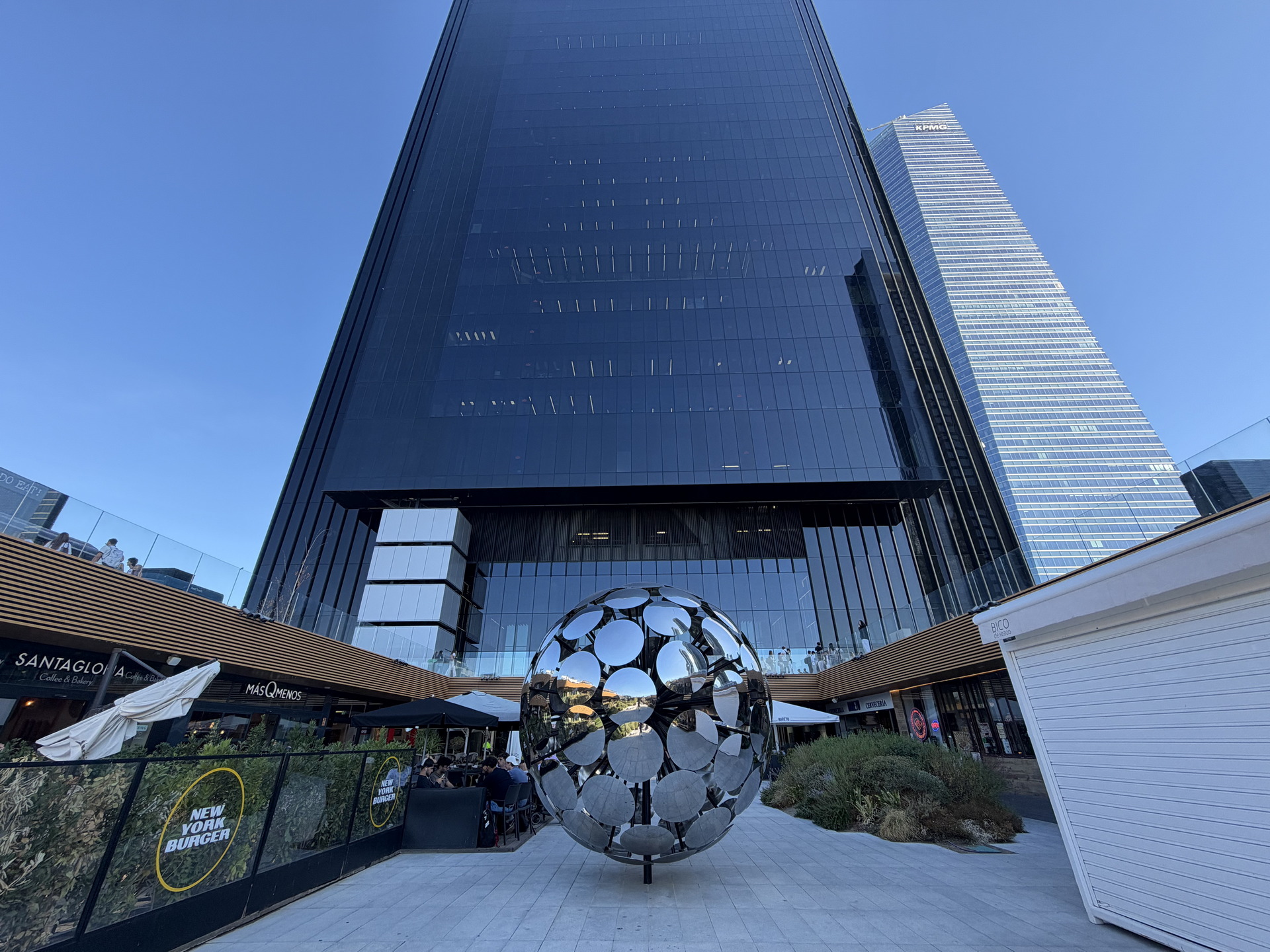 Rascacielos modernos con escultura esférica metálica en plaza urbana, cielo despejado y reflejos arquitectónicos.