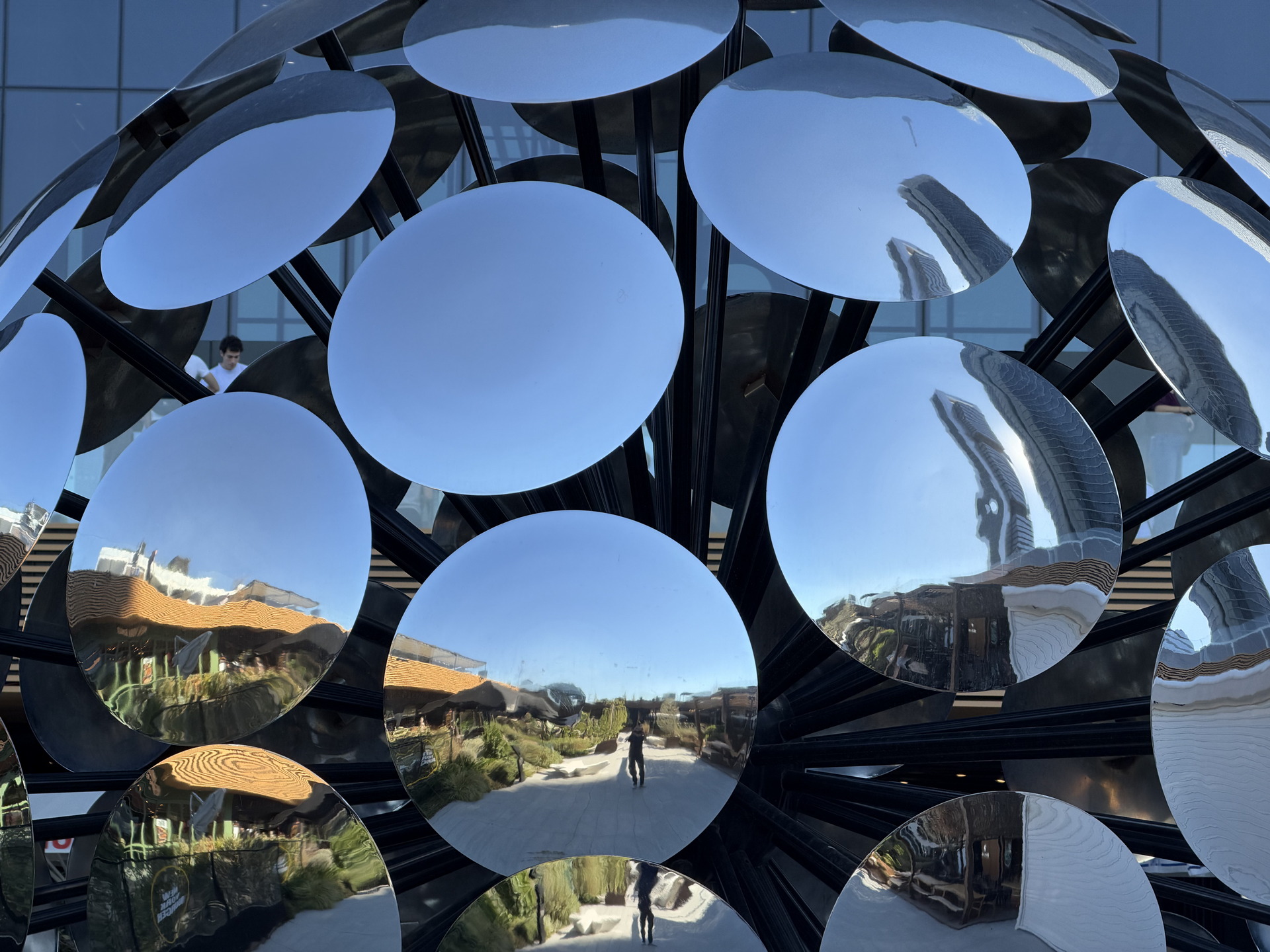 Escultura de círculos metálicos reflectantes con reflejos de edificios y cielo azul en el fondo urbano.