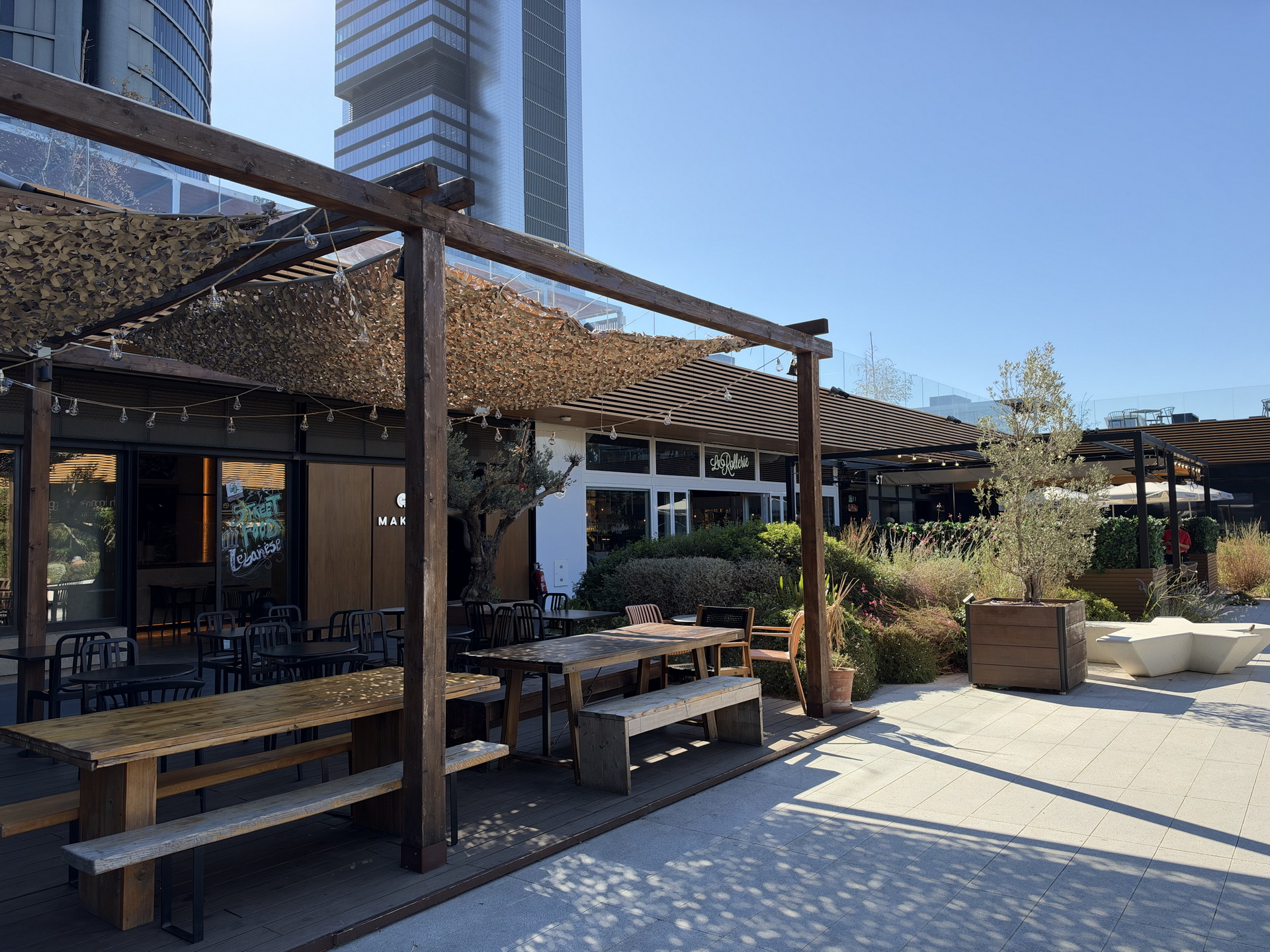 Terraza al aire libre de un restaurante moderno con mesas de madera y decoración vegetal bajo un cielo despejado.