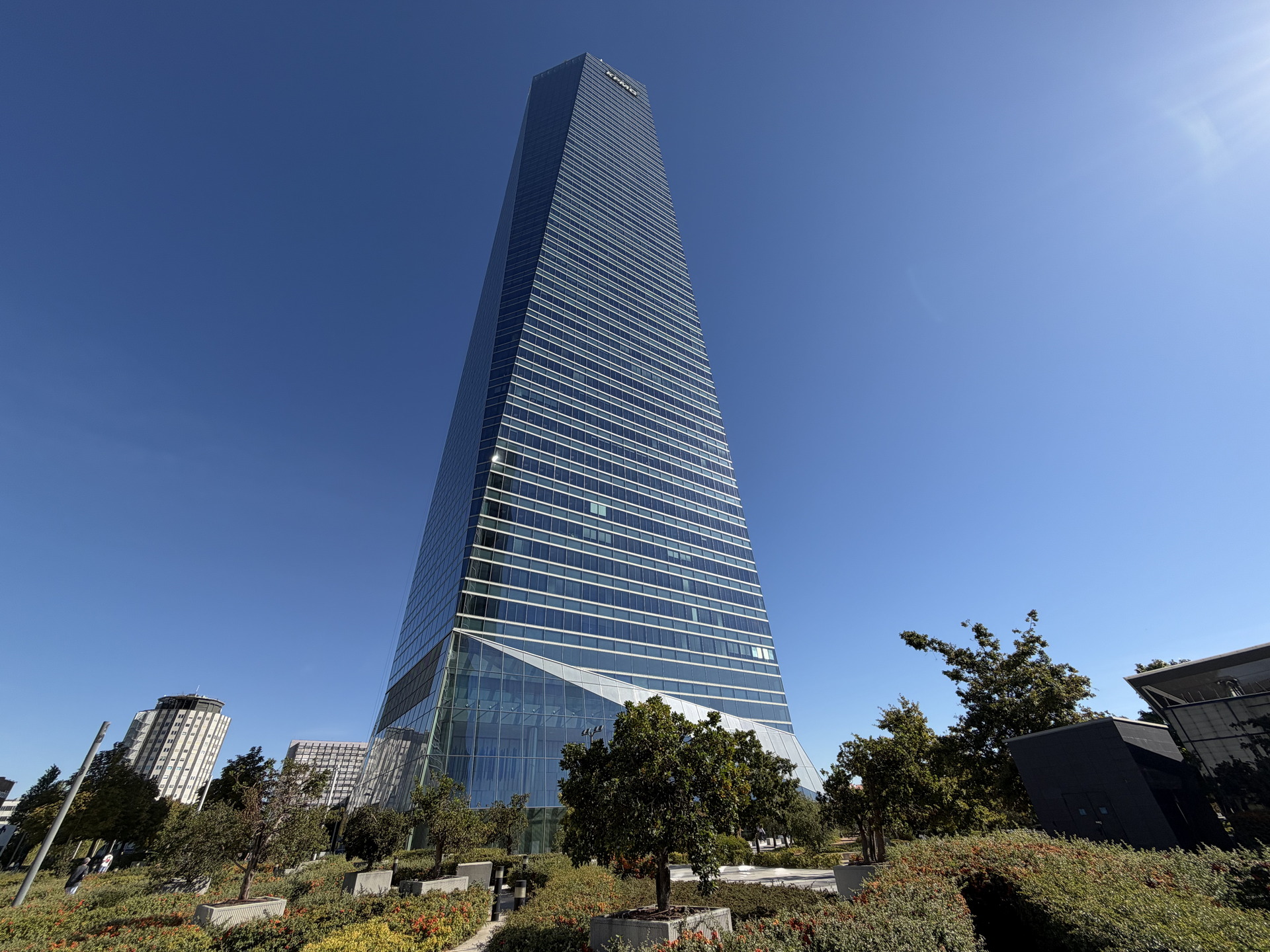 Rascacielos moderno reflejando el cielo azul claro, con jardines en primer plano y edificios menores alrededor.