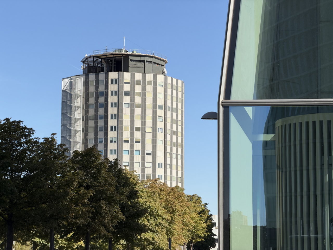 Edificio moderno con fachada de cristal y torre circular, rodeado de árboles bajo cielo azul.