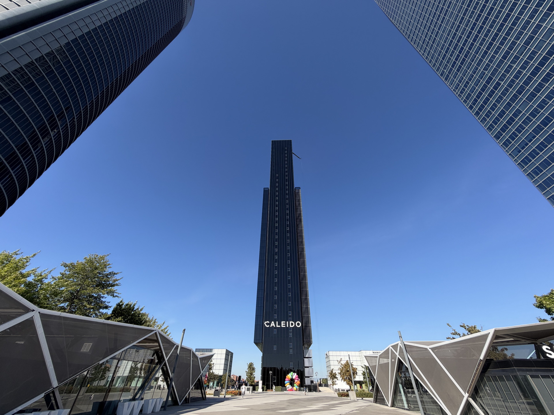 Edificio Caleido en Madrid con rascacielos y estructuras modernas en un cielo despejado azul.
