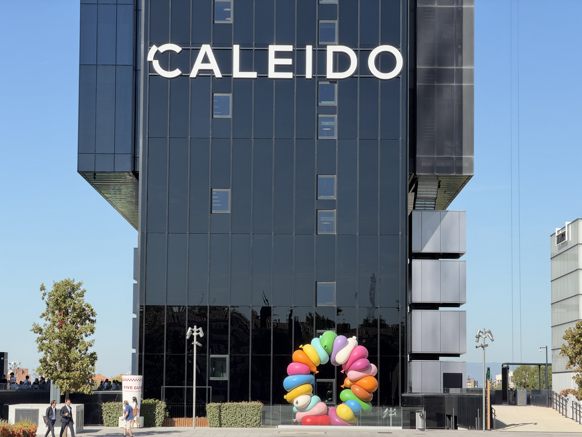 Edificio CALEIDO con escultura colorida frente a su fachada moderna en un día soleado.