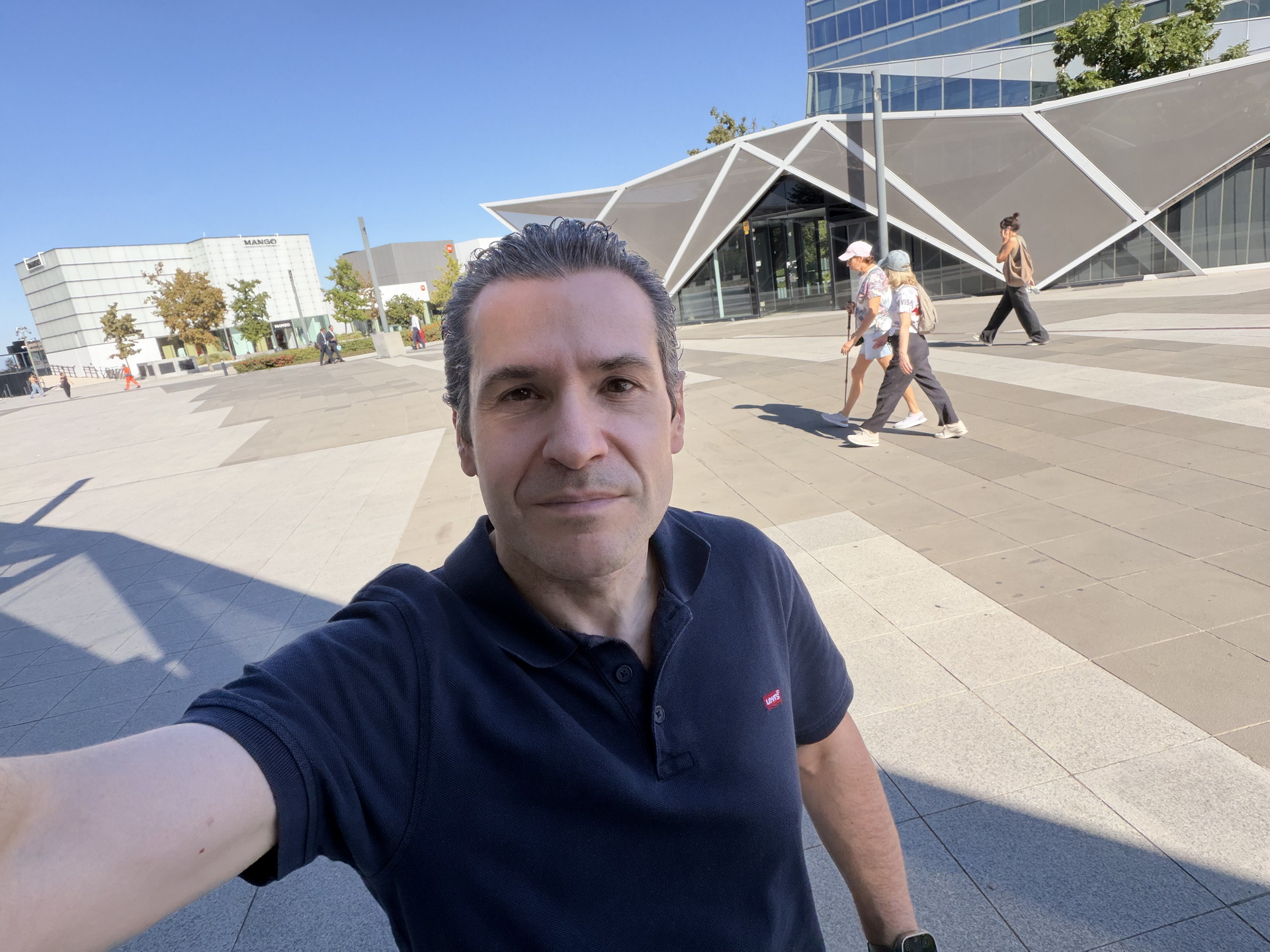 Hombre sonriente en una plaza moderna, con edificios contemporáneos y personas caminando al fondo, cielo despejado.