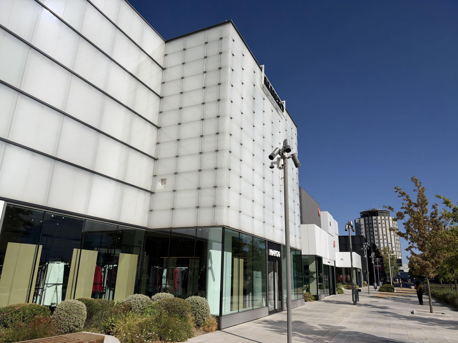 Centro comercial moderno con fachada blanca y escaparates en un día soleado. Árboles y arbustos en la acera.