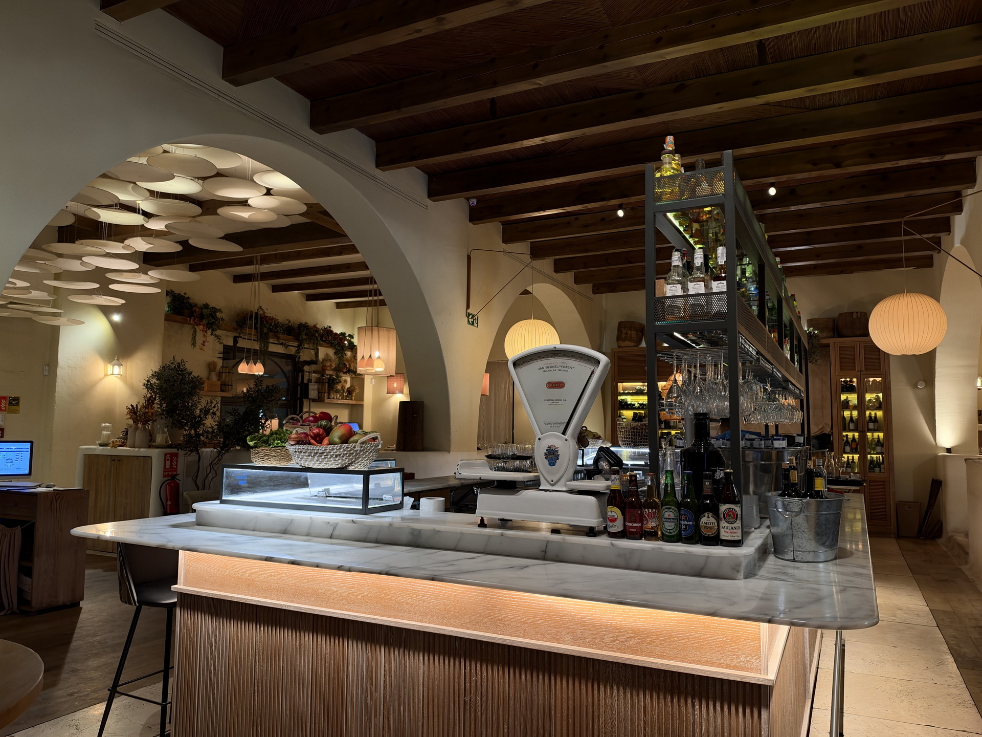 Interior de un restaurante acogedor con decoración moderna, barra iluminada y estantes con botellas y frutas.