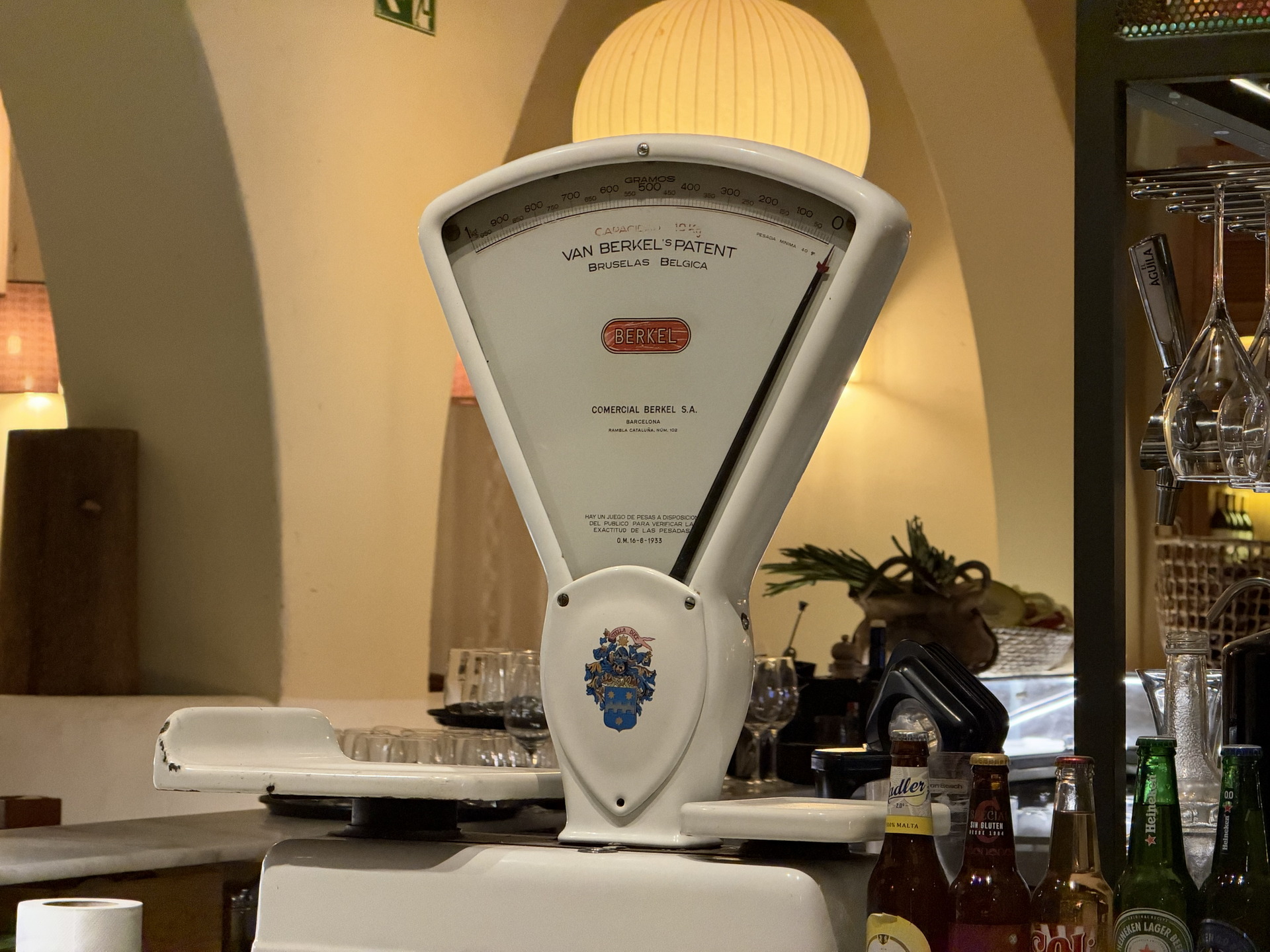 Báscula antigua Van Berkel blanca en un restaurante, rodeada de botellas y copas bajo una cálida iluminación.