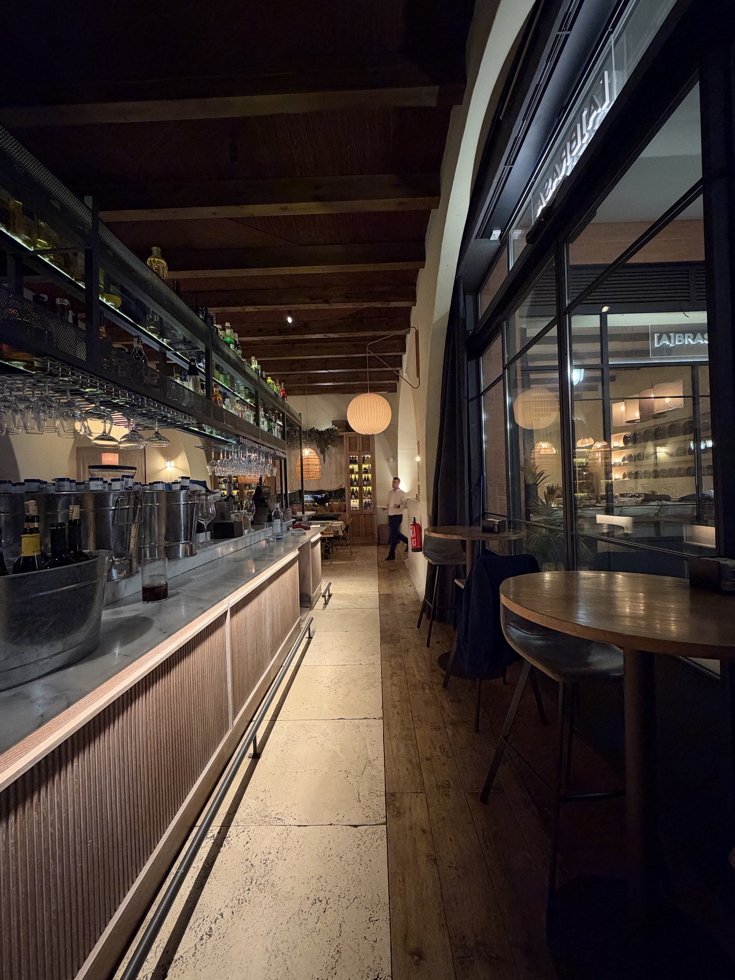 Interior acogedor de un bar moderno con iluminación suave, estantes de botellas y mesas junto a grandes ventanales.