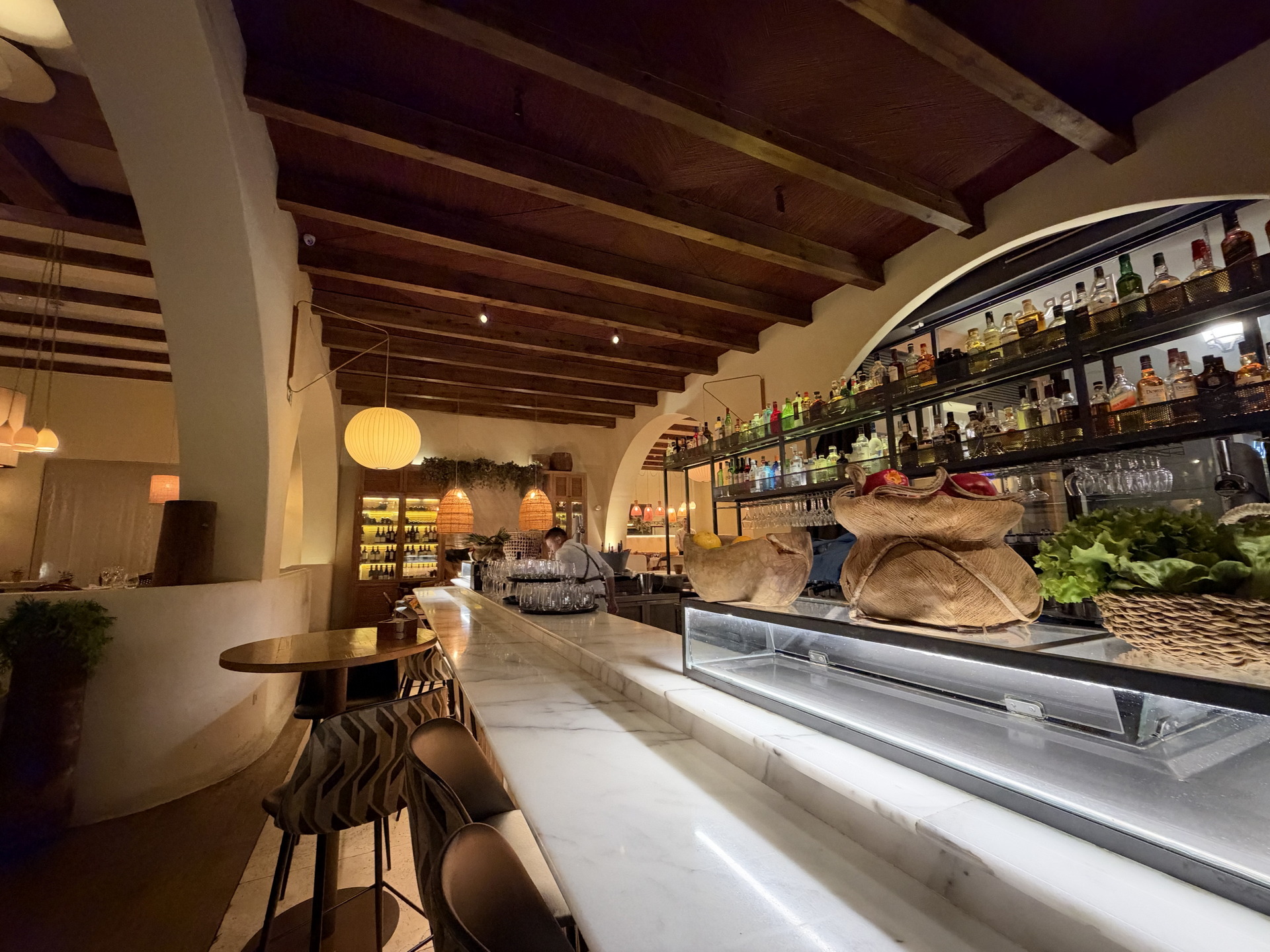 Bar y restaurante acogedor con decoración rústica, barra de mármol y estantes llenos de botellas iluminadas.
