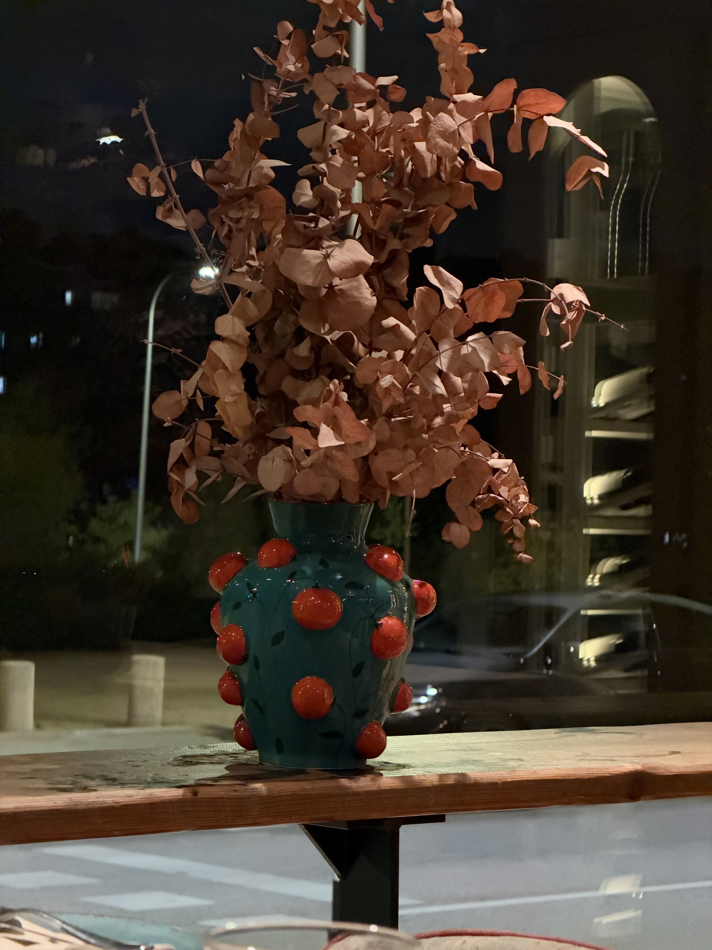 Jarrón azul con decoración roja y ramas secas, ubicado en una mesa junto a una ventana nocturna.