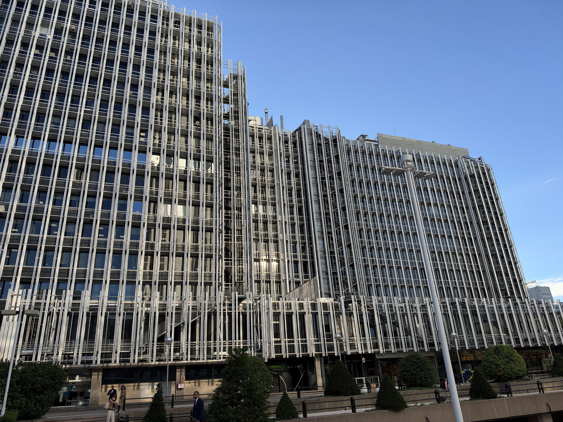 Edificio contemporáneo con fachada de acero y vidrio bajo un cielo azul claro, rodeado de árboles y personas caminando.