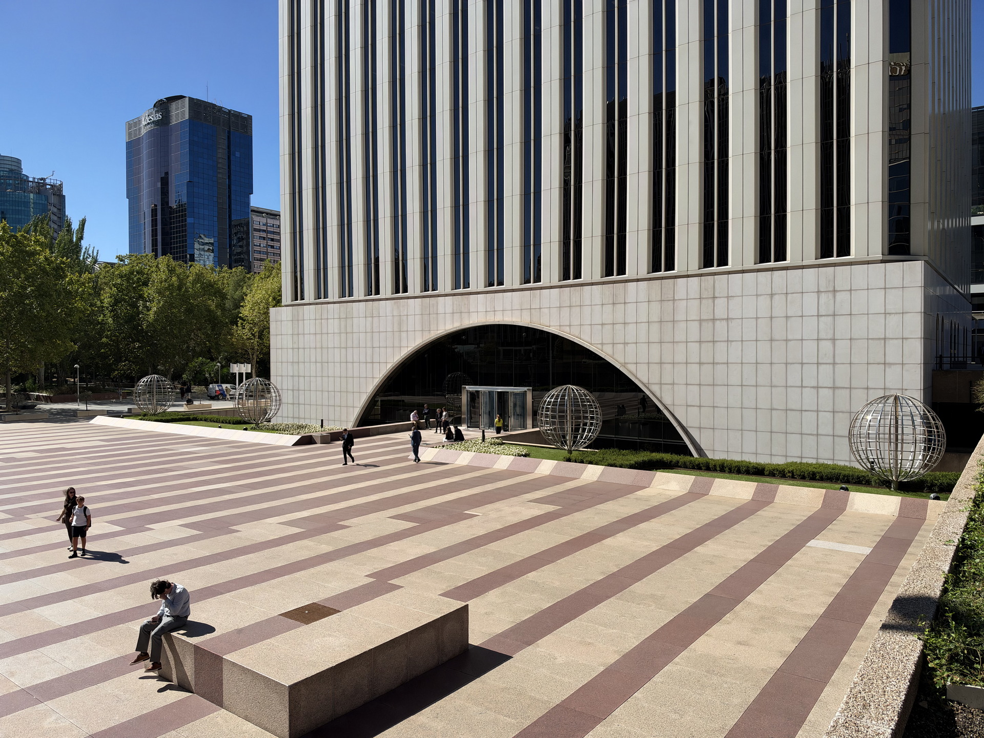 Plaza moderna frente a edificio de oficinas con esculturas esféricas y peatones en día soleado.