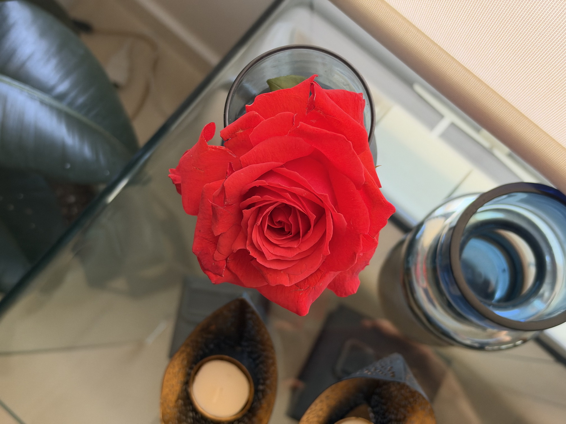 Rosa roja en un florero sobre mesa de vidrio junto a vela y decoraciones modernas.