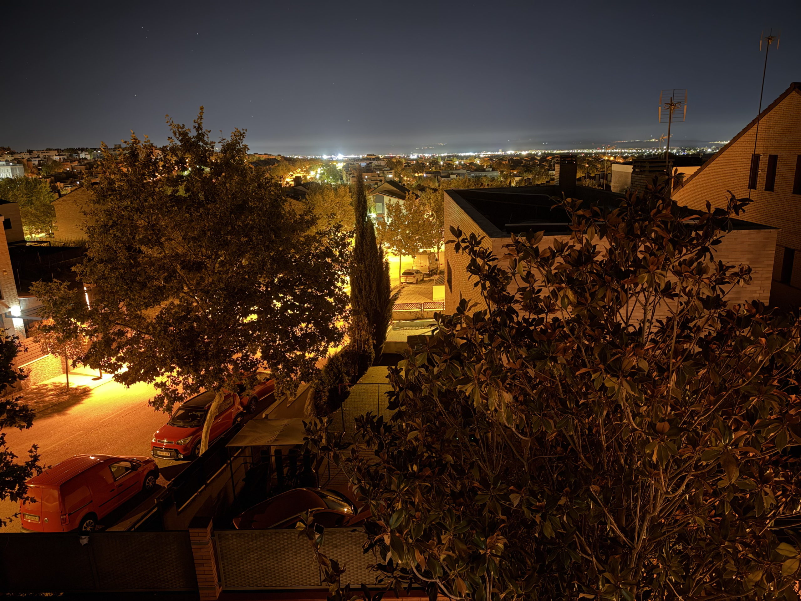 Vista nocturna de una calle iluminada por farolas, con árboles y casas en el horizonte bajo un cielo estrellado.