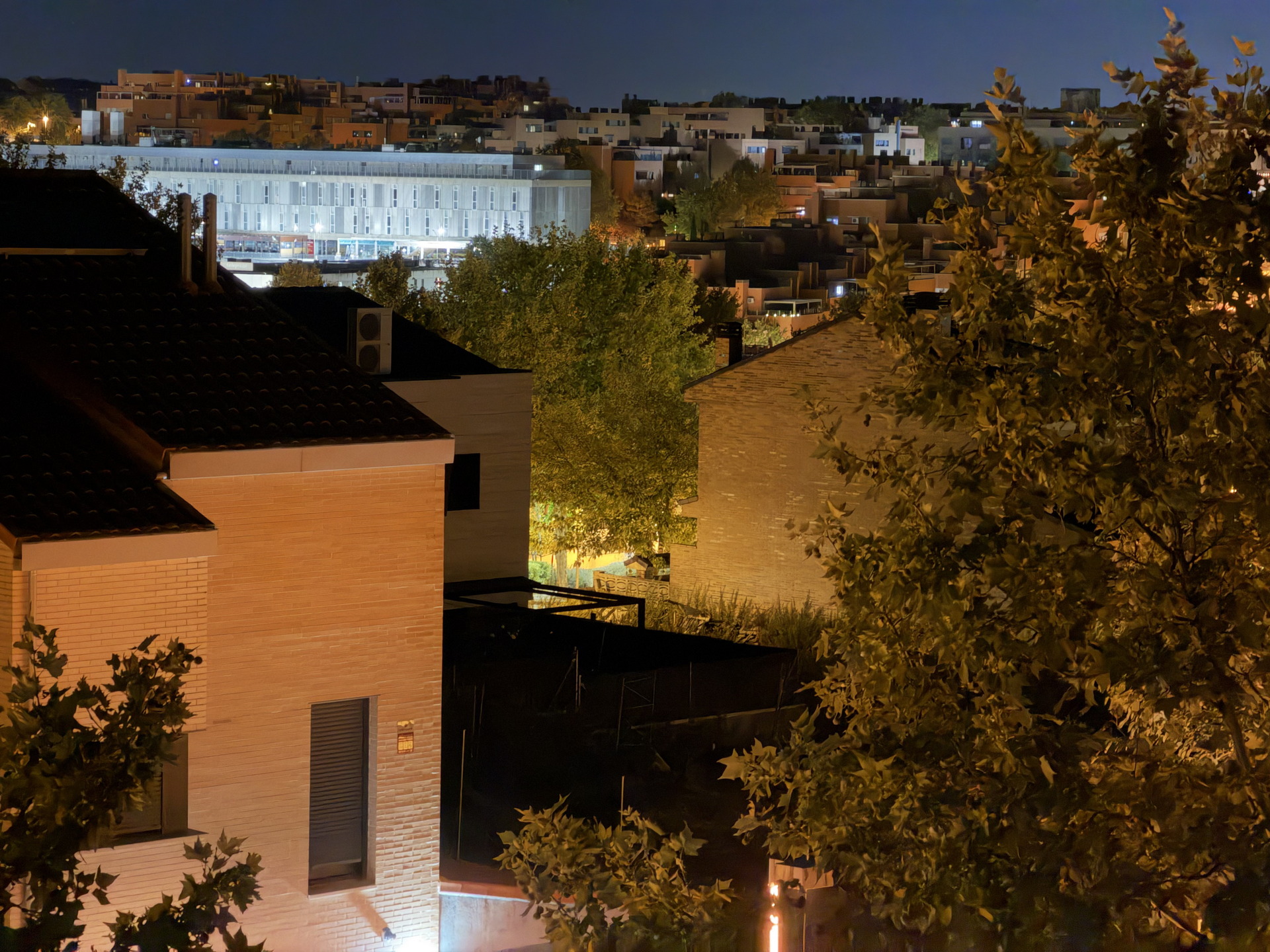 Vista nocturna de un barrio residencial con casas y árboles iluminados, fondo de ciudad bajo cielo claro.