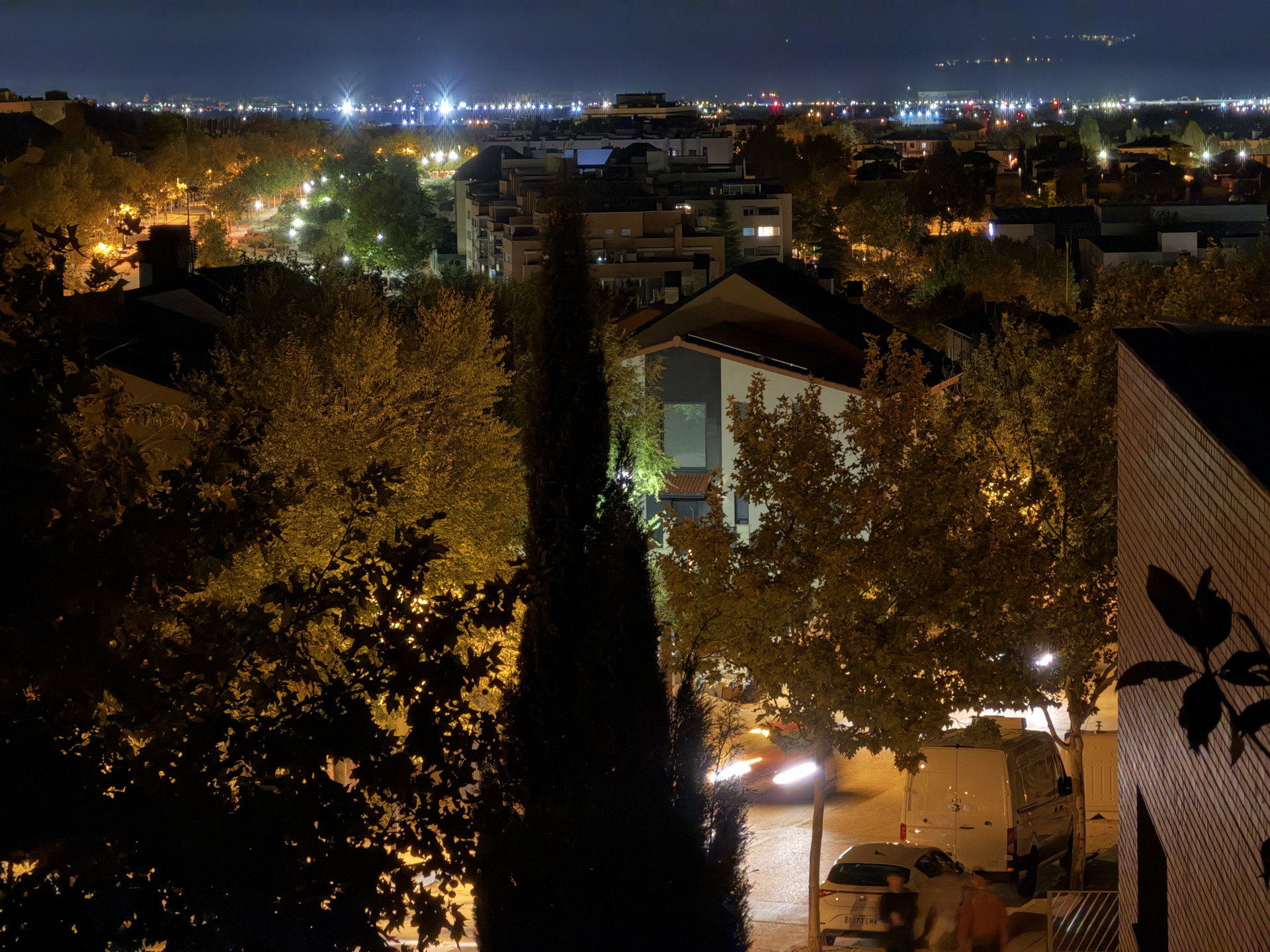 Vista nocturna de una ciudad iluminada desde las alturas, con árboles, coches y edificios bajo un cielo estrellado.