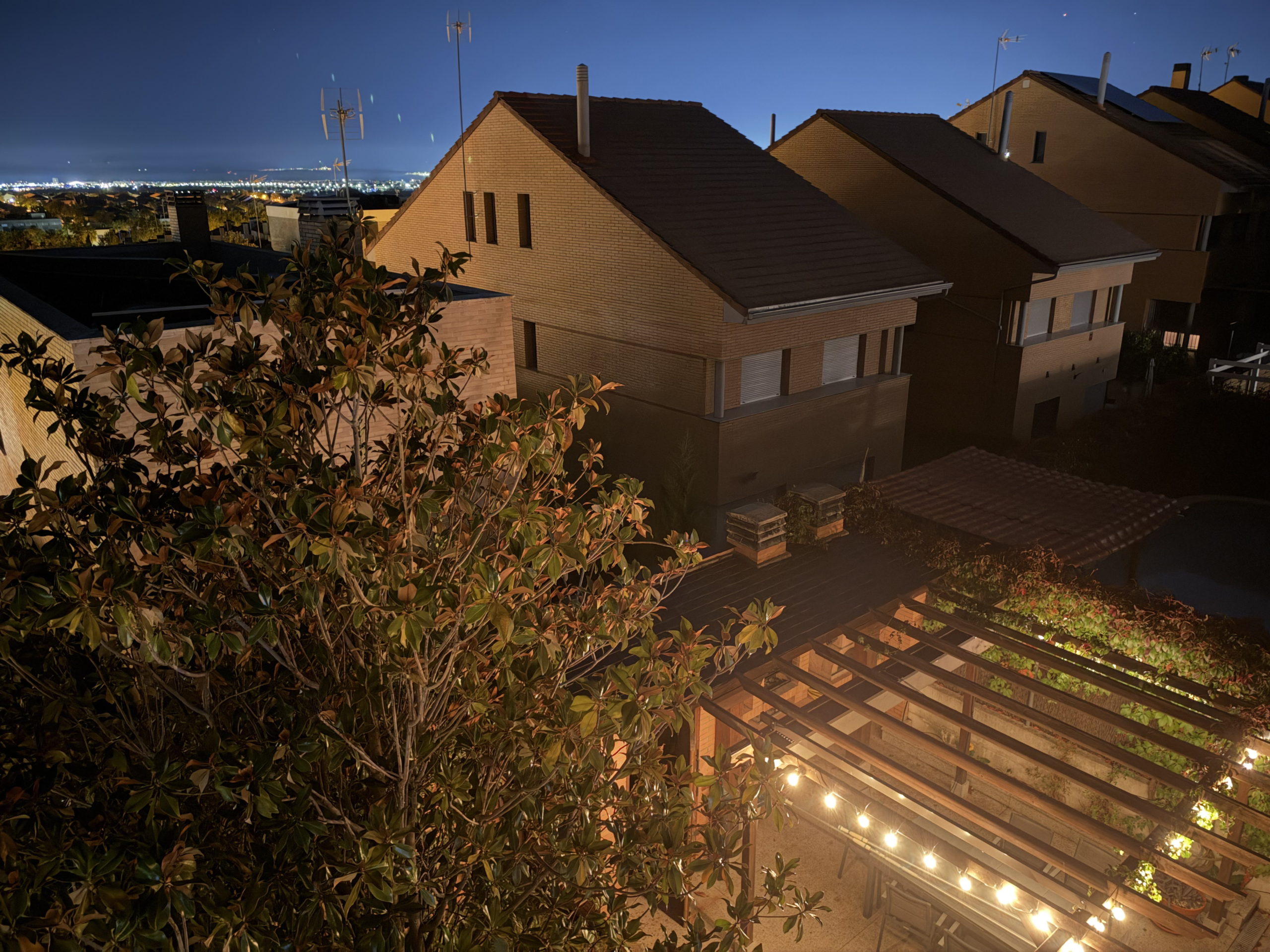 Terraza iluminada de noche con pérgola de luces y casas de fondo. Vista urbana tranquila y moderna.