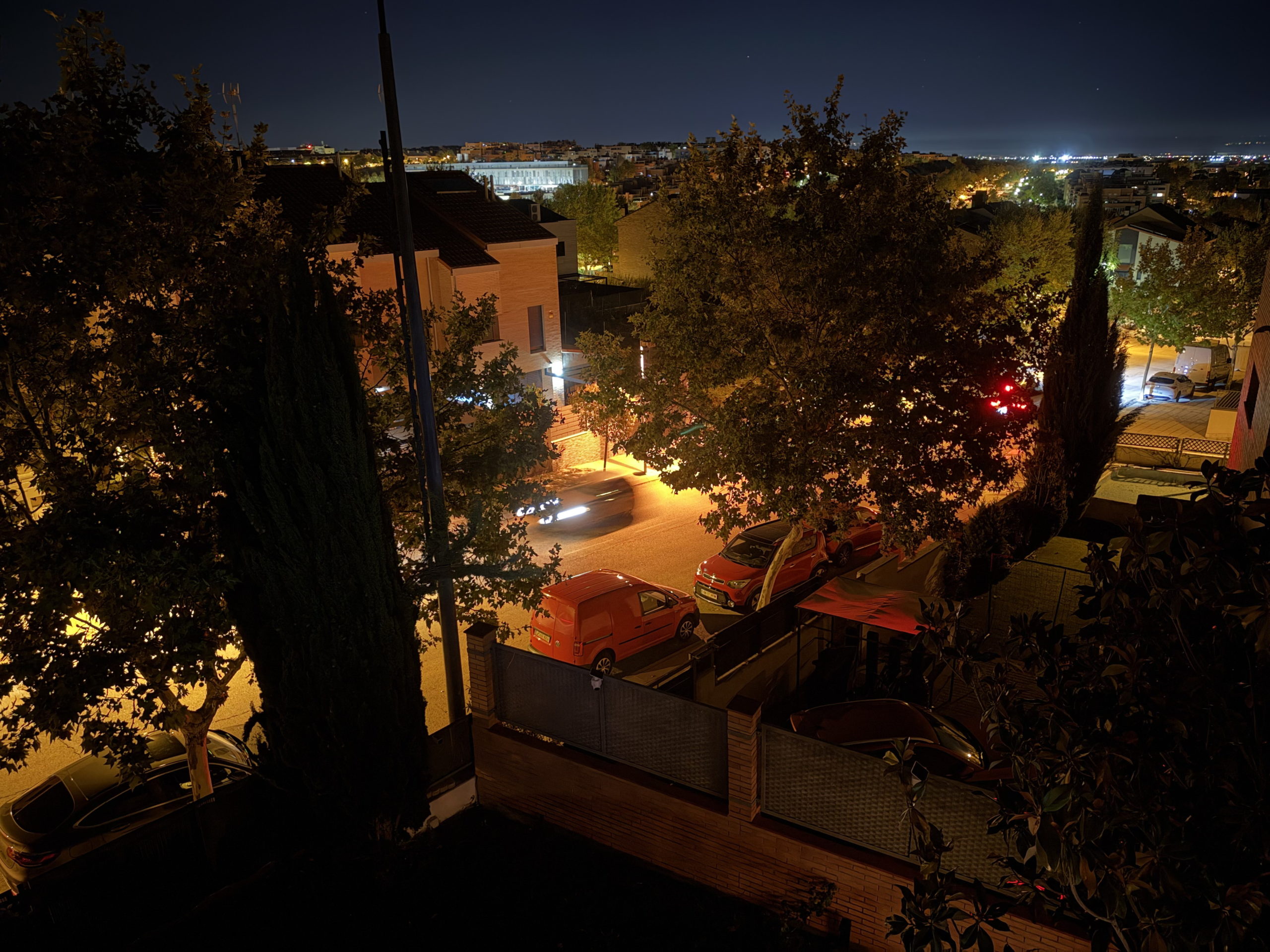 Calle iluminada de noche con coches estacionados y árboles, vista desde un balcón.
