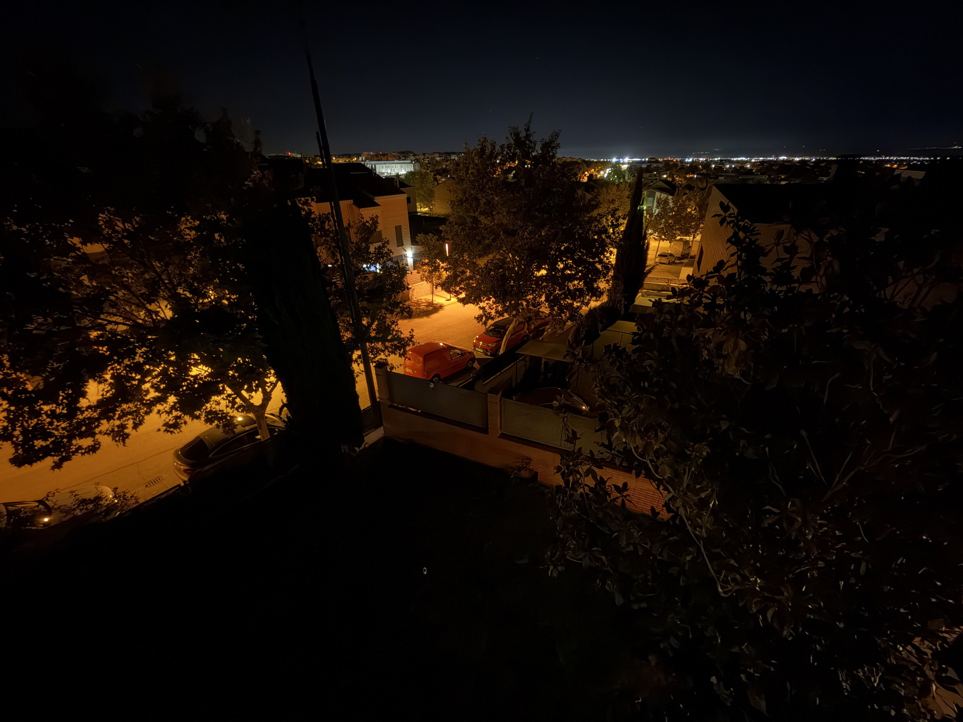 Vista nocturna de una calle iluminada por farolas, rodeada de árboles y automóviles estacionados.