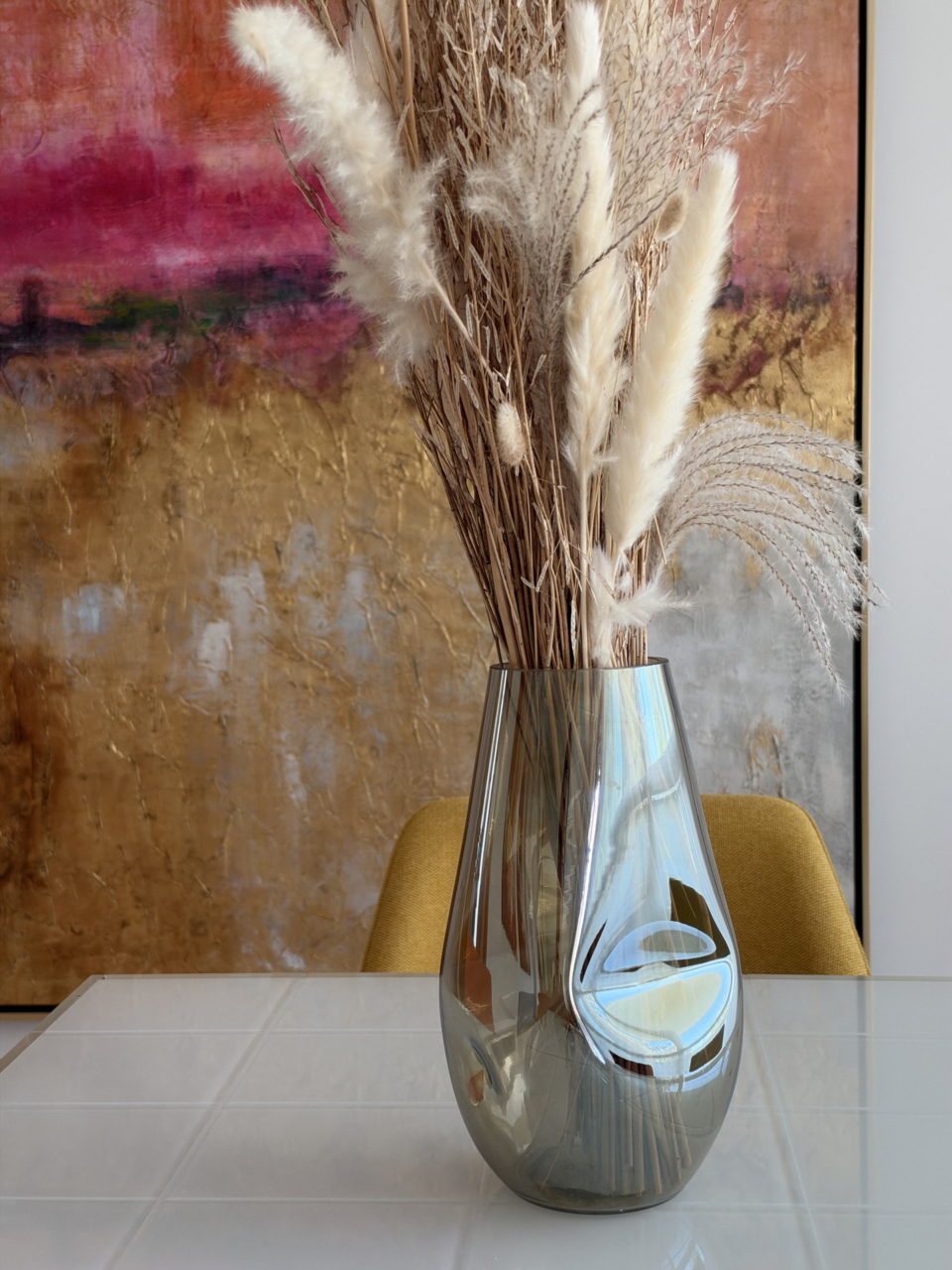Jarrón de vidrio con flores secas sobre mesa blanca, fondo de arte abstracto colorido en sala moderna.