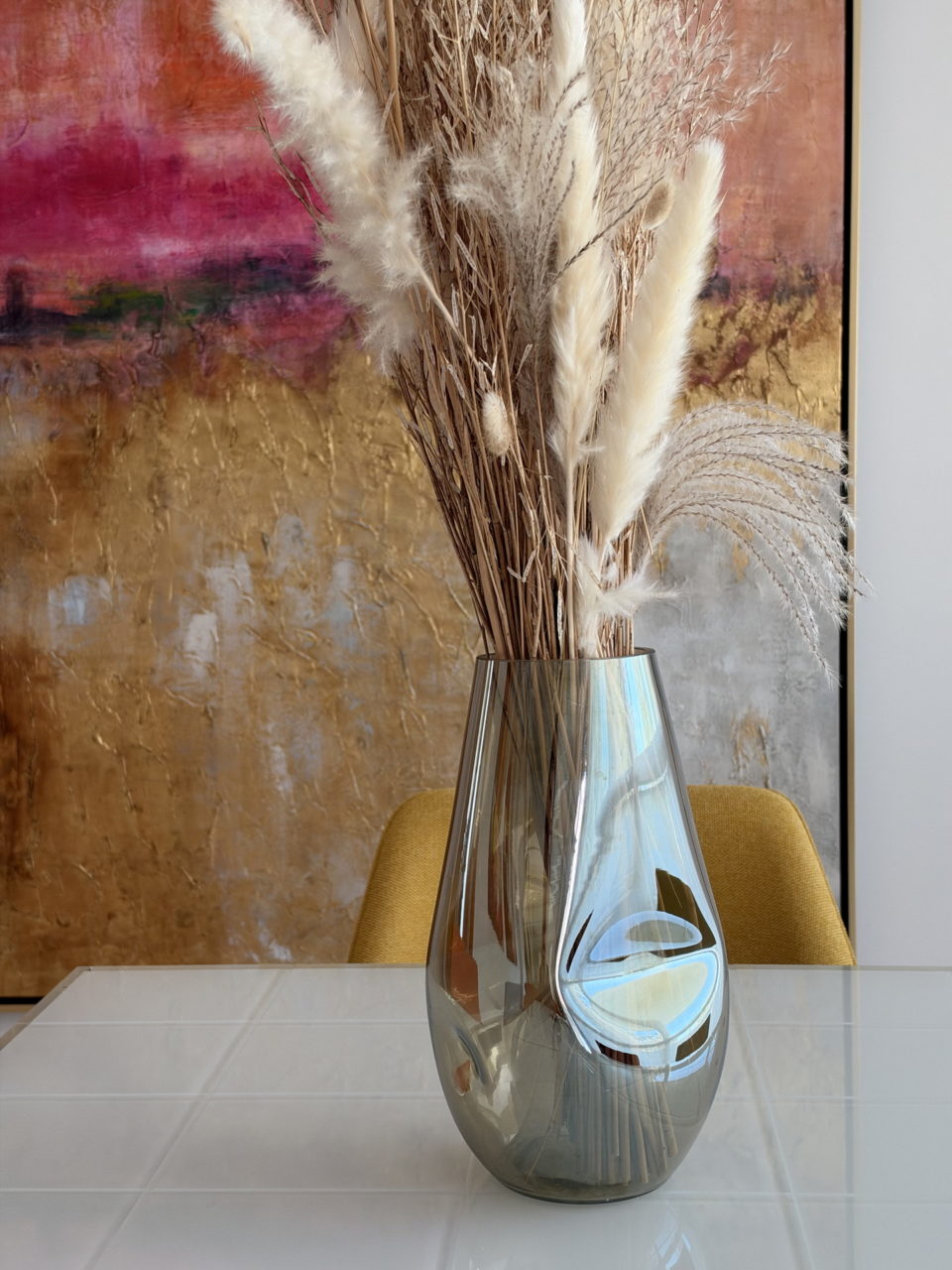 Jarrón de cristal con flores secas sobre mesa blanca, fondo de arte abstracto en tonos cálidos.