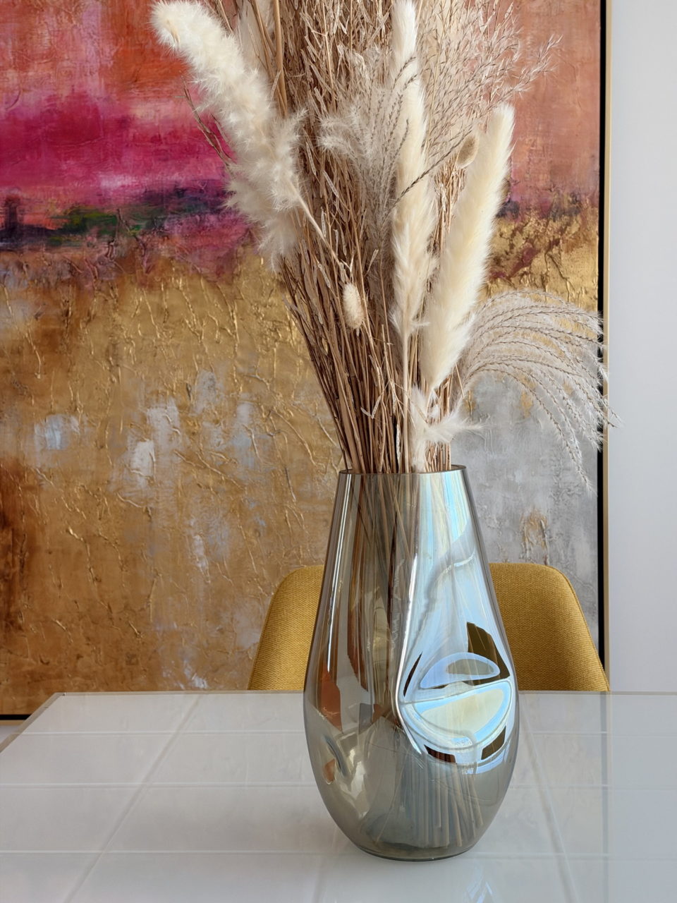 Florero de vidrio con hierbas decorativas sobre mesa blanca, fondo de arte abstracto en tonos dorados y rosados.